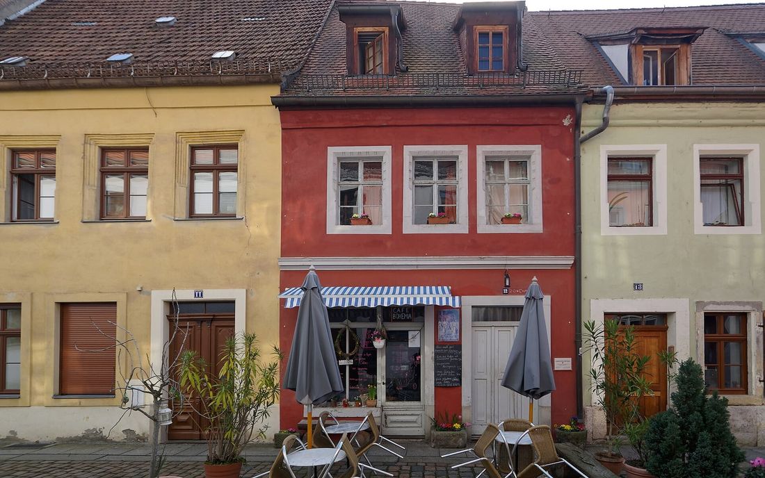 Fachada de casas antigas com um pequeno café no térreo, toldo de lona listrada azul e branca, ombrelones e mesas na calçada