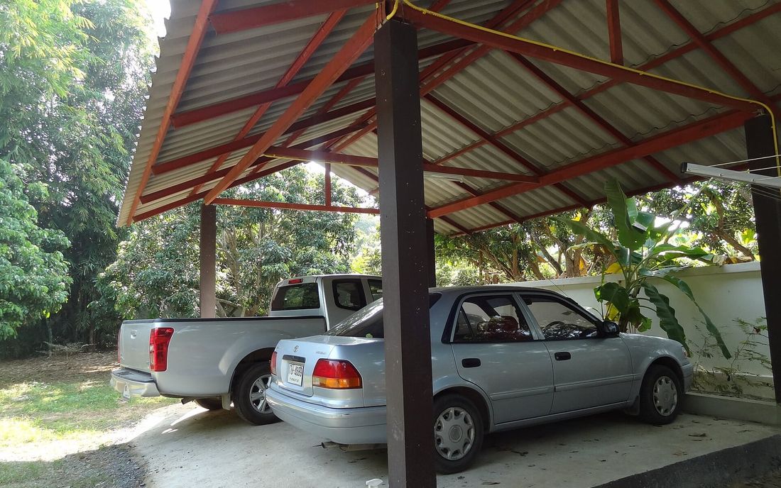 Garagem coberta com estrutura metálica vermelha e telhas onduladas, com uma caminhonete e um carro estacionados
