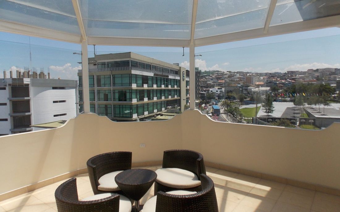 Varanda no alto de um prédio com telhado transparente, poltronas e mesa, com vista para a cidade